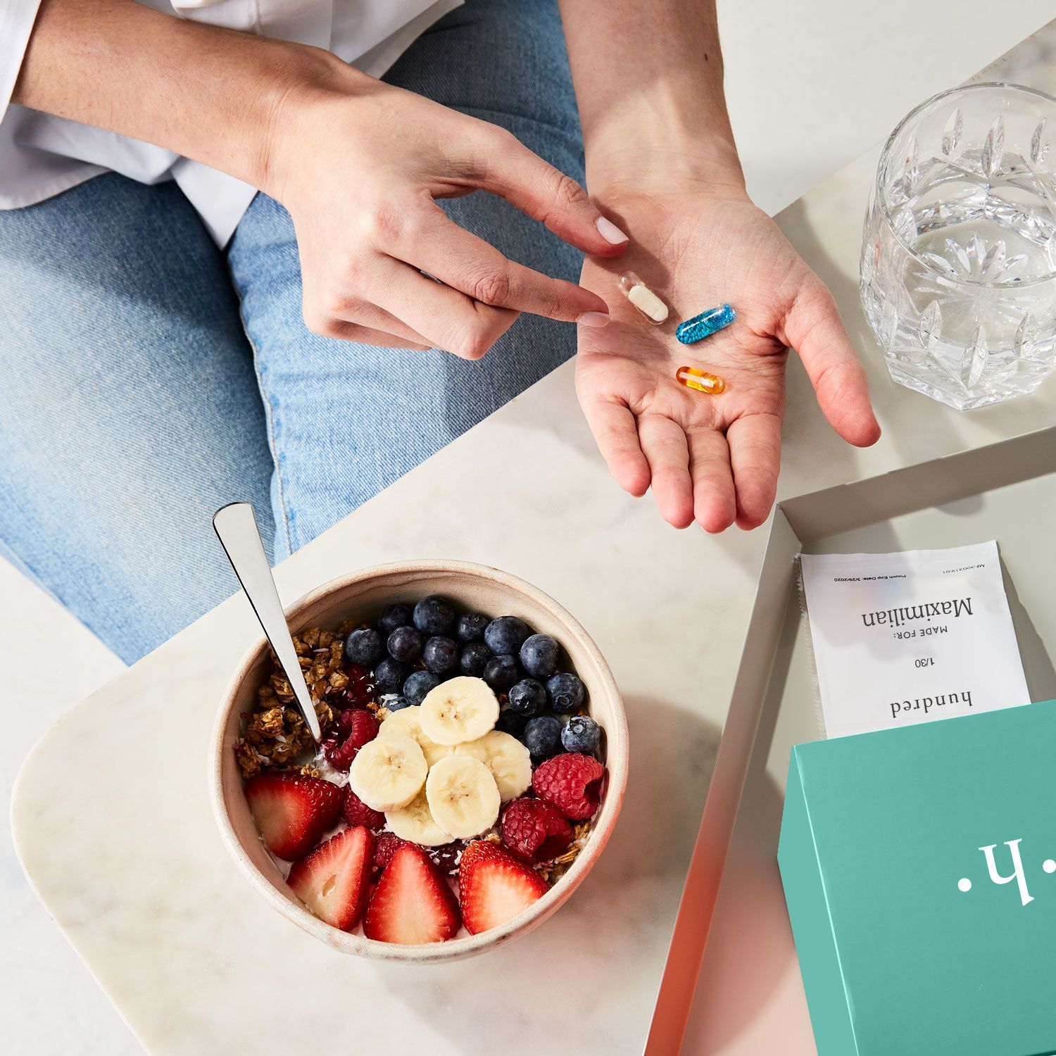 Woman holds pills and supplement vitamins in here hand over a marble table with a green box and fruit cereal bowl on countertop