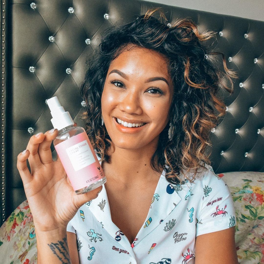 A beautiful young woman in a fun patterned shirt holds a Brandless rosewater facial toner in her floral bedroom