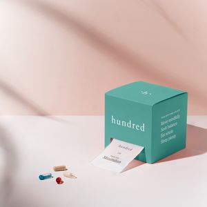 vitamins and supplements lay on a white table in front of a millennial pink backdrop with a hundred box that has a personalized pill pack coming out of it